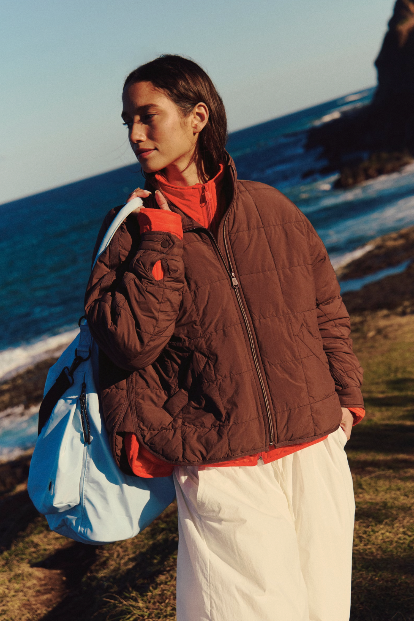 A woman standing outdoors by the ocean wearing a brown quilted puffer jacket over a bright orange top and white pants, with a light blue bag slung over her shoulder.