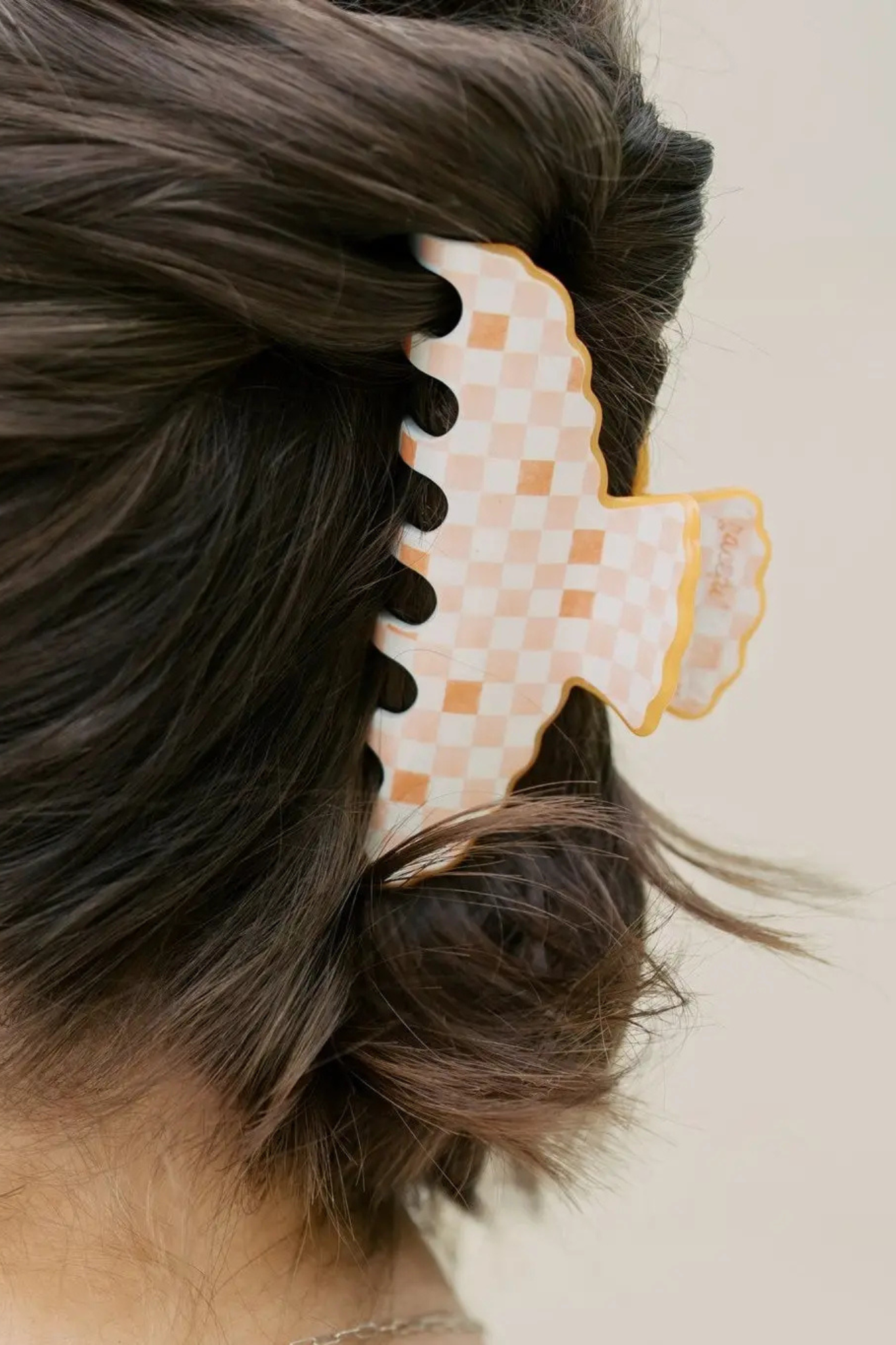 A close-up of a person's dark brown hair secured with a large claw-style hair clip. The clip has a white and light orange checkered pattern with a yellow border, shown against a plain light beige background.