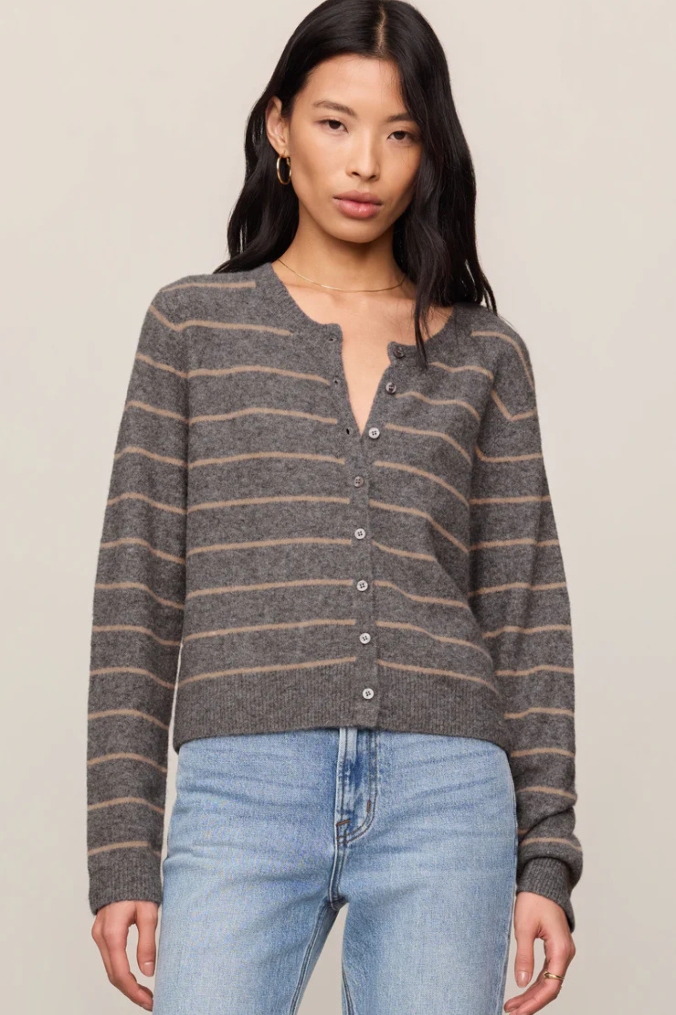 A woman wearing a gray and brown striped knit henley cardigan with light blue jeans, photographed against a plain beige studio background.