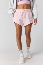 A model wearing light pink sweat shorts with a drawstring waist, styled with a gray hoodie, pink sports bra, white socks, and white sneakers against a light gray studio background.