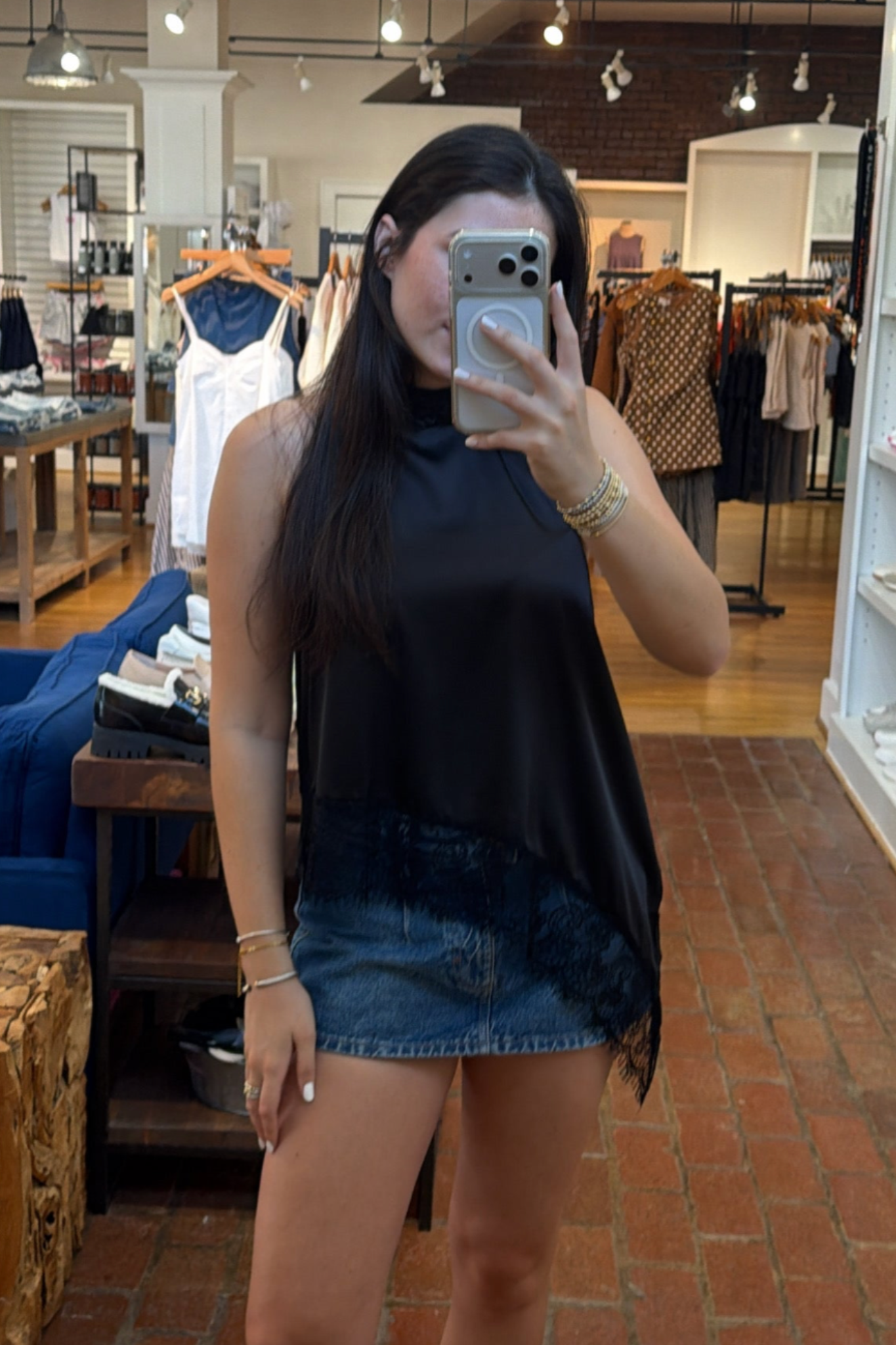 A woman takes a mirror selfie in a clothing boutique, wearing a black satin halter top with a sheer lace hem and a denim mini skirt.