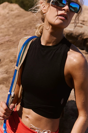 A woman outdoors wearing a black sleeveless crop top, red shorts, sunglasses, and carrying a blue hydration pack on her shoulder.