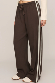 A person wearing dark brown wide-leg sweatpants with double white side stripes, a drawstring waist, and white slip-on shoes, shown against a plain light gray studio background.