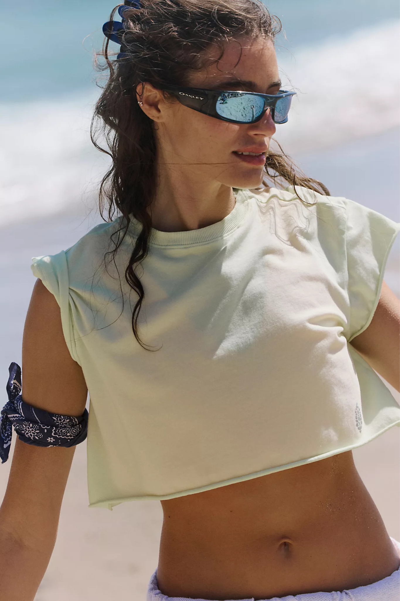 A woman on a beach wearing a pale yellow-green cropped sleeveless top and dark sunglasses with blue mirrored lenses.