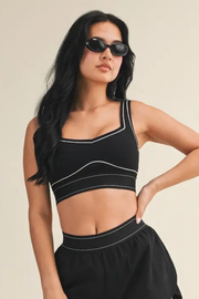 A woman modeling a black crop top with white contrast piping and a matching black skirt against a solid beige background.