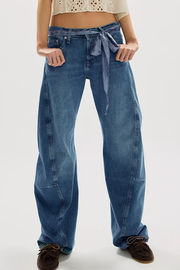 A person wearing wide-leg blue denim jeans with a tie waist, paired with brown shoes, shown from the waist down against a light gray studio background.
