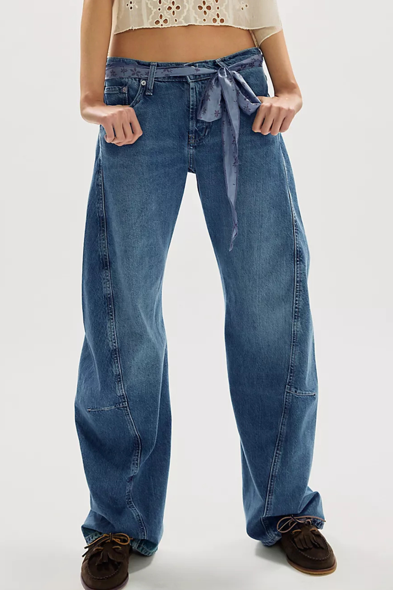 A person wearing wide-leg blue denim jeans with a tie waist, paired with brown shoes, shown from the waist down against a light gray studio background.