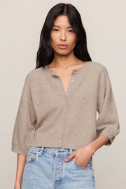 A woman wearing a light taupe knit cardigan with three-quarter sleeves and a buttoned split neckline, paired with light blue jeans, against a solid light beige background.