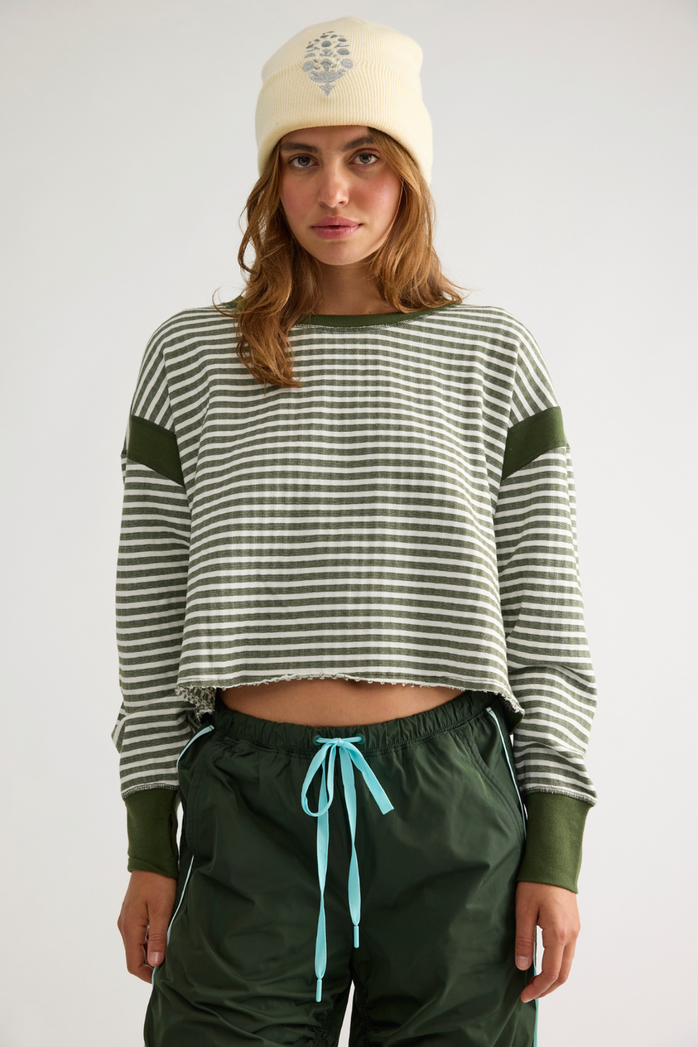 A woman wearing a cropped long-sleeve knit top with green and white horizontal stripes and solid green ribbed cuffs and shoulders, paired with dark green drawstring pants and a cream beanie, against a light gray studio background.