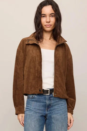A woman modeling a brown suede cropped jacket over a white top and blue jeans, photographed against a plain light beige studio background.