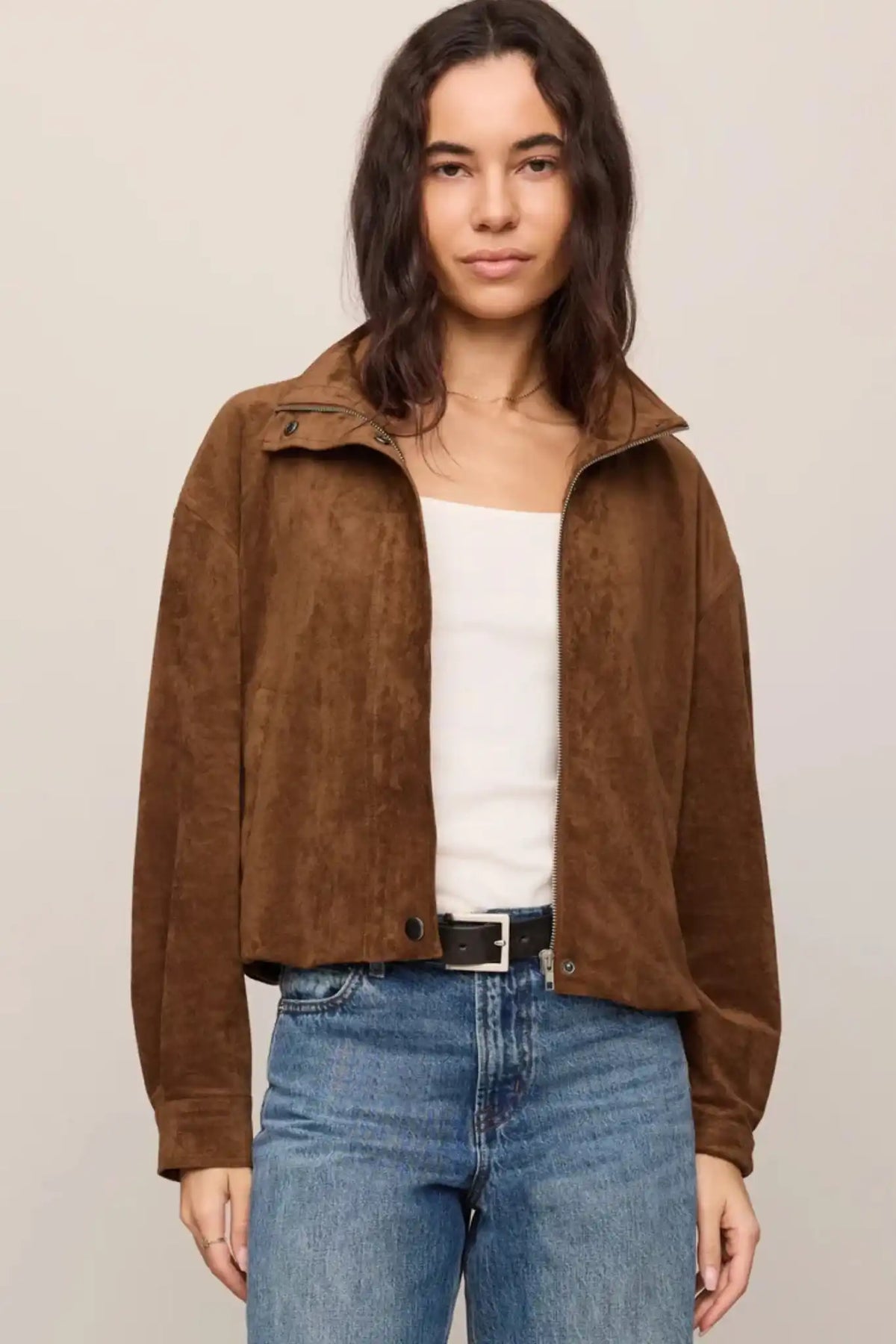A woman modeling a brown suede cropped jacket over a white top and blue jeans, photographed against a plain light beige studio background.