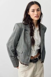 A woman modeling a cropped gray plaid blazer with thin brown and red windowpane lines, worn over a white button-up shirt and cream pants, against a light gray studio background.