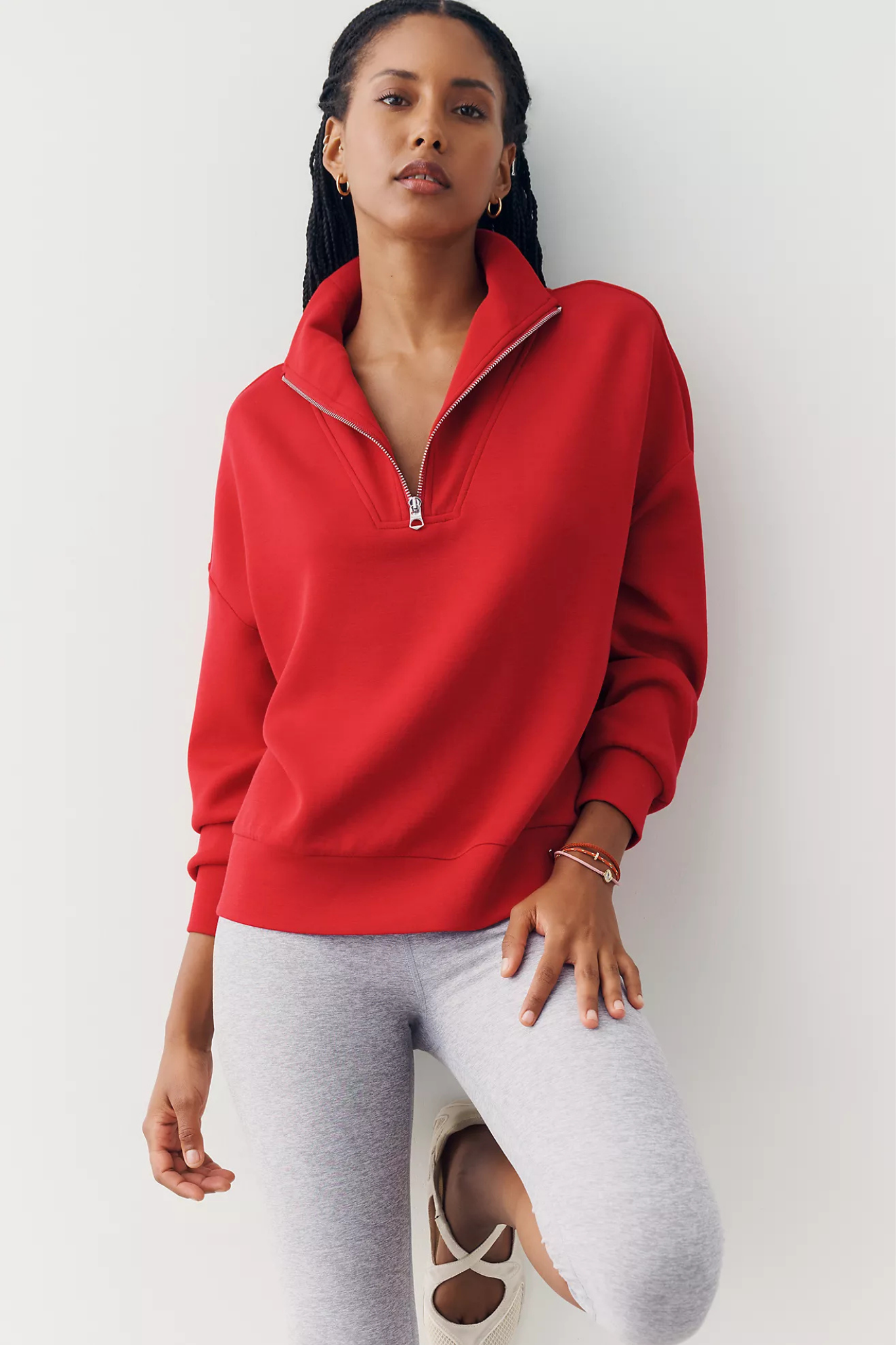 A woman wearing a bright red half-zip sweatshirt and light gray leggings, posing against a clean white wall.