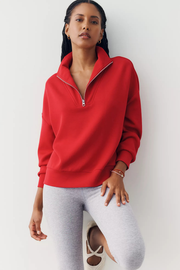 A woman wearing a bright red half-zip sweatshirt and light gray leggings, posing against a clean white wall.