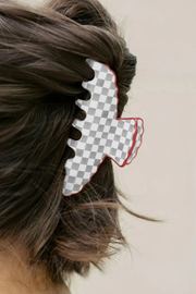 A close-up side view of a person's hair secured with a large hair claw clip. The clip has a gray and white checkerboard pattern with a red outline, set against a plain light beige background.