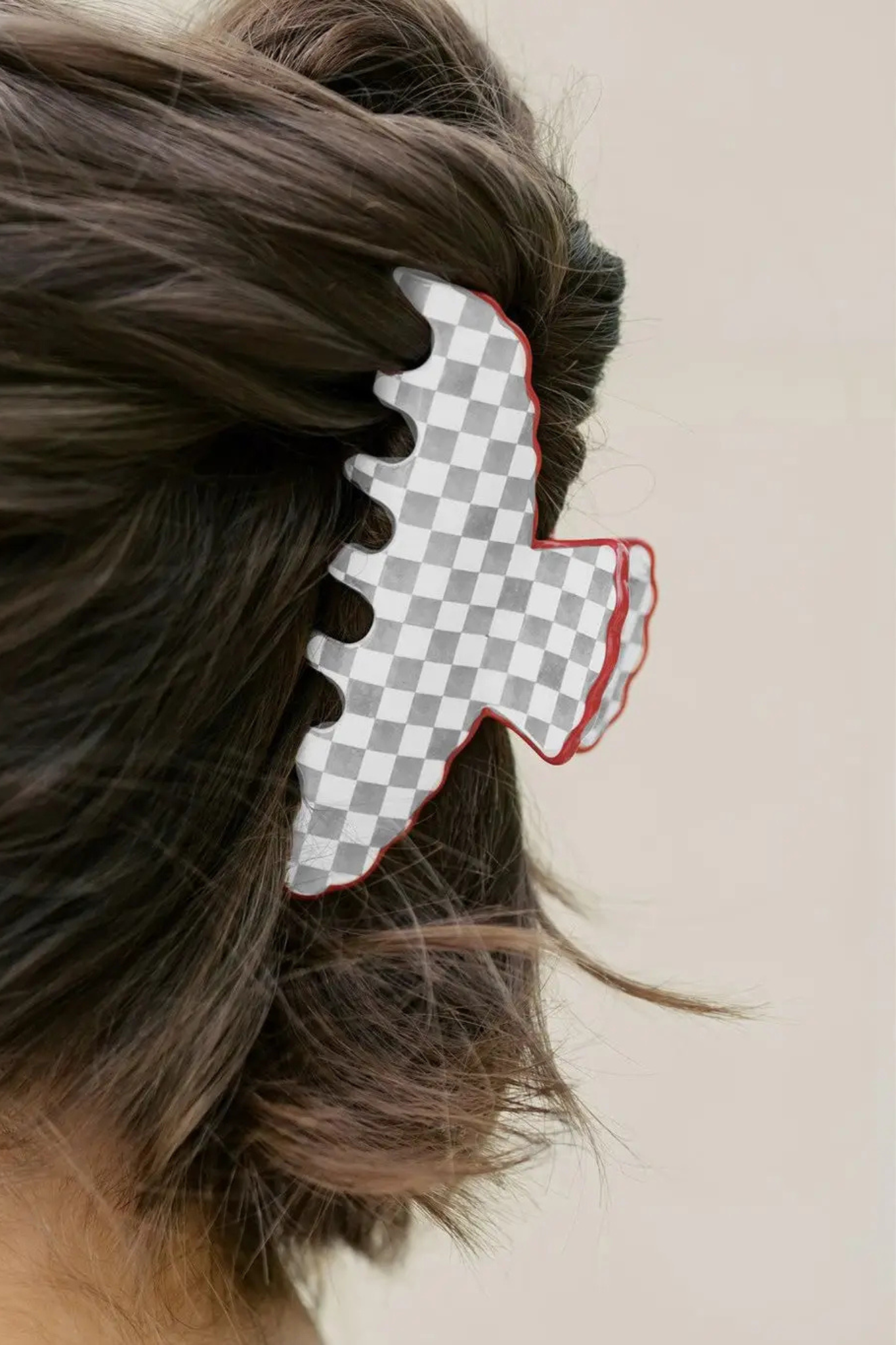 A close-up side view of a person's hair secured with a large hair claw clip. The clip has a gray and white checkerboard pattern with a red outline, set against a plain light beige background.