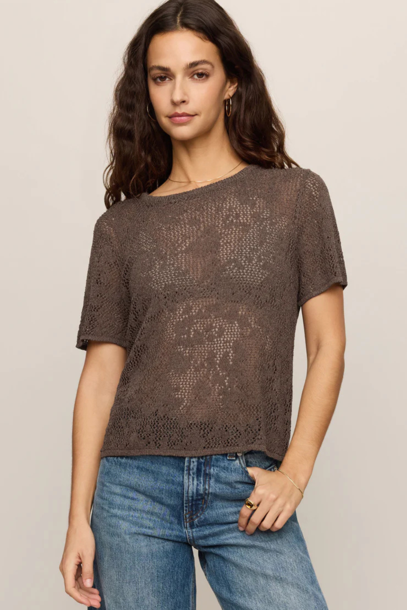 A woman wearing a brown short-sleeve knit top with an open lace-like pattern, paired with blue jeans, against a plain light beige studio background.
