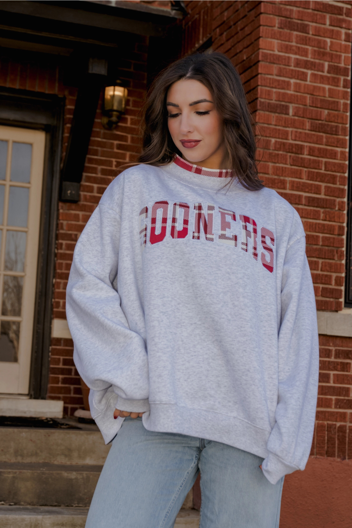 A woman wearing a light gray heather sweatshirt with 'RODERS' printed in red across the chest, paired with light blue jeans, standing in front of a red brick building.