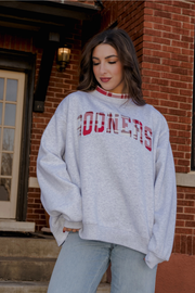A woman wearing a light gray heather sweatshirt with 'RODERS' printed in red across the chest, paired with light blue jeans, standing in front of a red brick building.