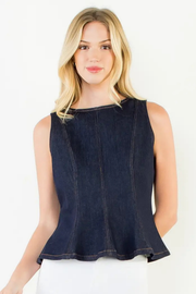 A blonde woman wearing a dark indigo denim sleeveless top with a peplum hem, paired with white bottoms, photographed against a plain white studio background.