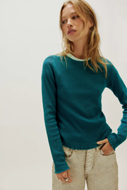 A woman wearing a teal long-sleeve knit sweater with a light green inner collar and beige pants, photographed against a plain light gray studio background.