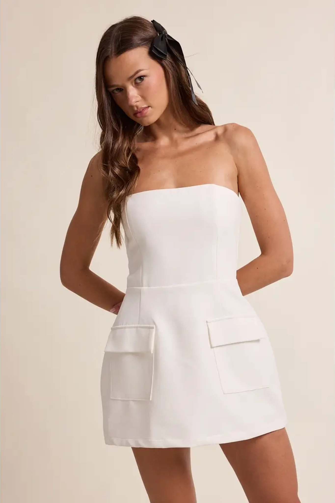A woman modeling a strapless white mini dress with large front patch pockets, photographed against a plain warm beige studio background.