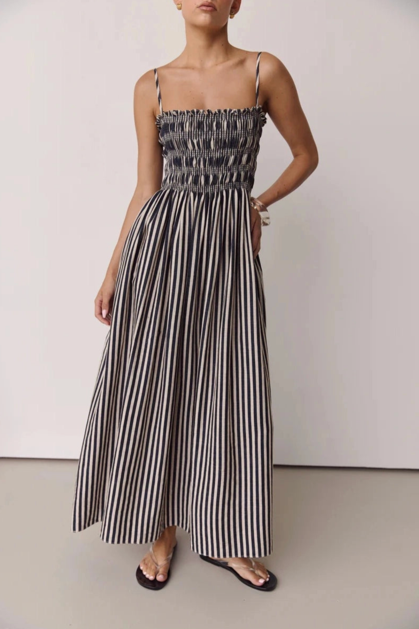 A woman modeling a sleeveless maxi dress with thin straps, a smocked bodice, and wide-leg striped pants in black and off-white, paired with black flat sandals against a plain studio background.