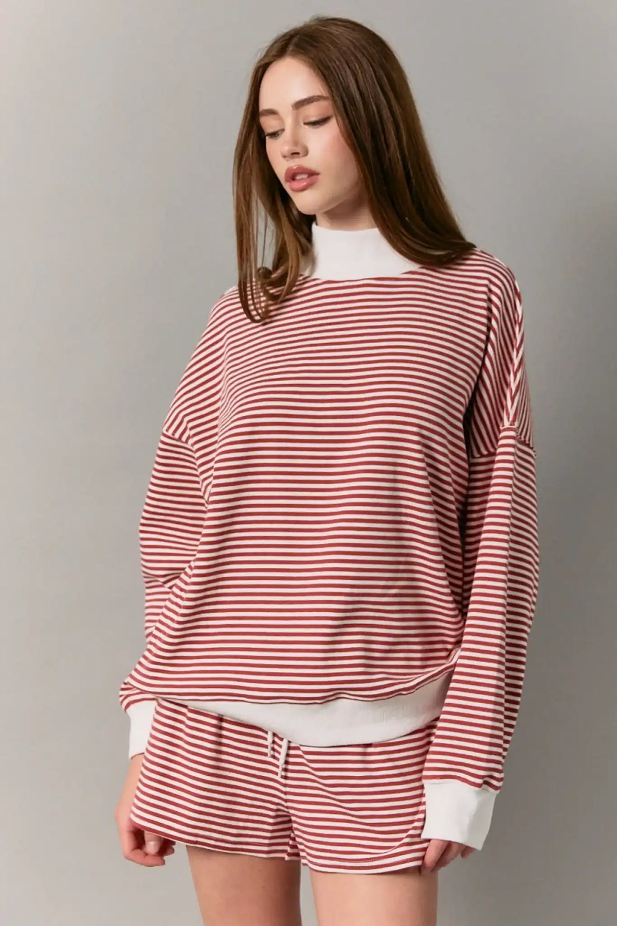A woman wearing a matching red and white striped lounge set, featuring a long-sleeve top and shorts, posed against a neutral gray studio background.
