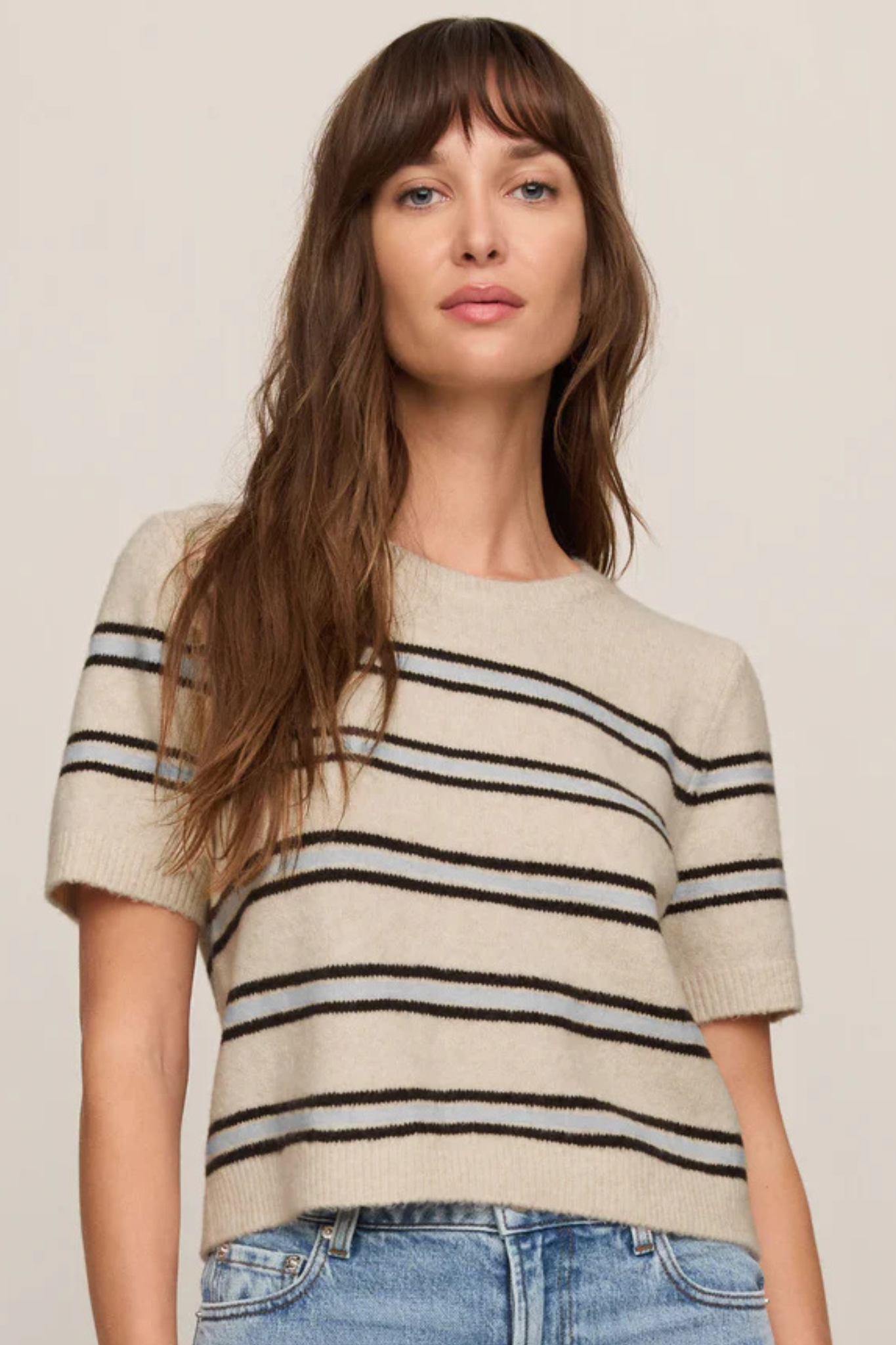 A woman wearing a short-sleeve knit sweater with horizontal black and gray stripes on a cream base, paired with blue jeans, against a plain light beige background.