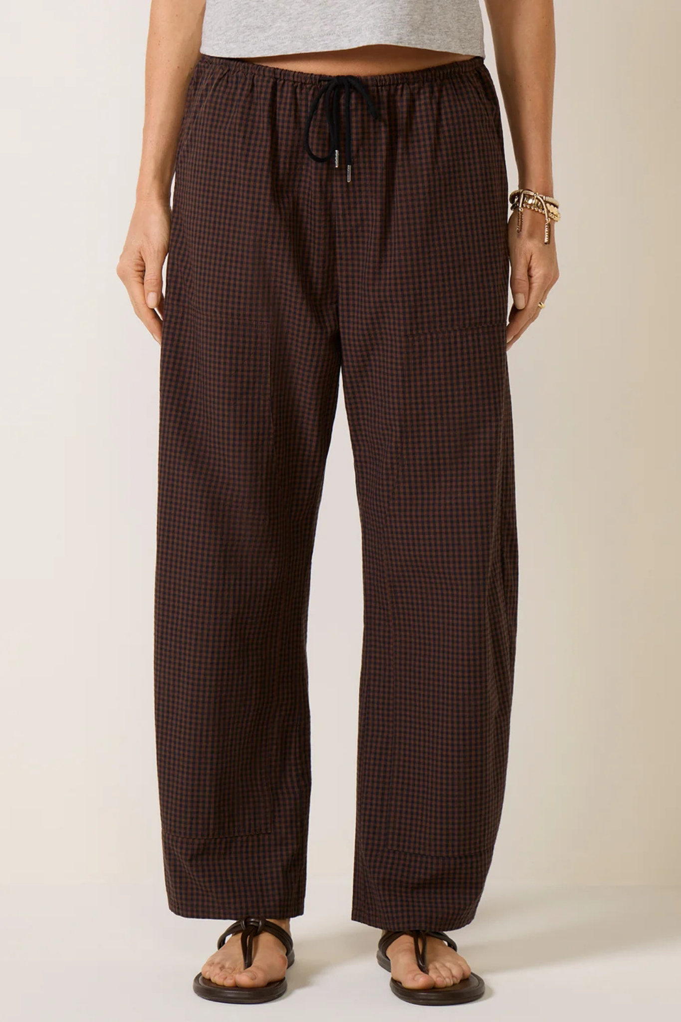 A person wearing dark brown and black checkered wide-leg pants with a drawstring waist, paired with brown sandals, shown against a plain beige studio background.