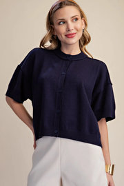 A woman wearing a navy blue short-sleeve button-up knit top and white pants, standing against a plain beige studio background.