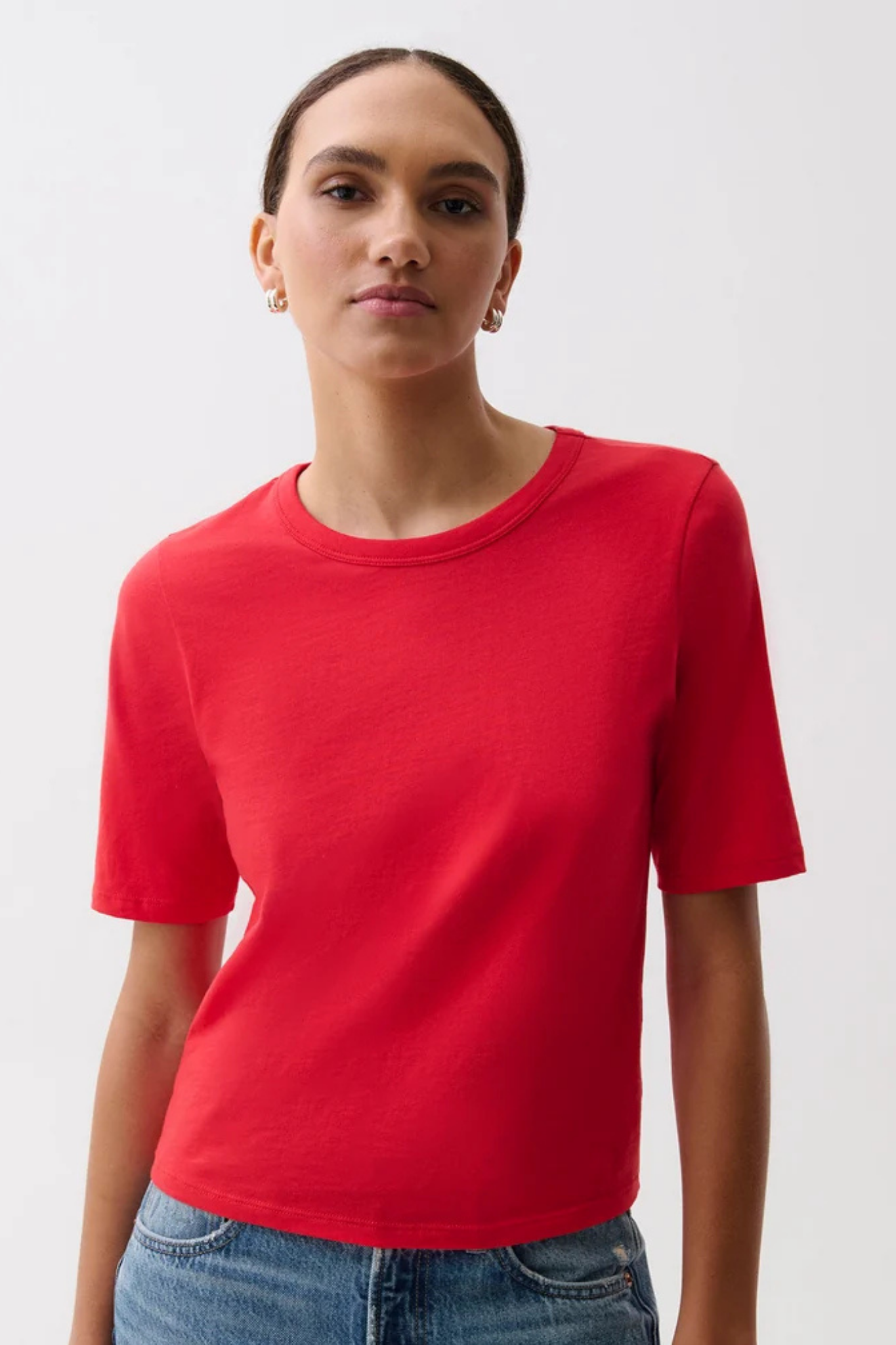 A woman wearing a bright red short-sleeve crewneck t-shirt and blue jeans, photographed against a clean white studio background.