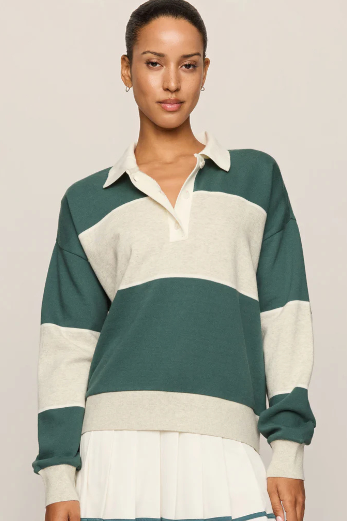 A woman wearing a long-sleeve rugby-style sweater with horizontal stripes in dark green and cream, paired with a white pleated skirt, against a plain light beige studio background.