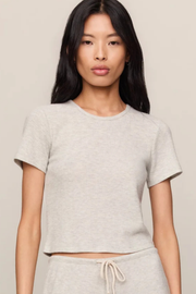 A woman modeling a light heather gray short-sleeve crop top and matching drawstring sweatpants against a plain beige studio background.
