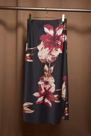 A dark navy midi skirt with a large red and cream floral print, hanging from a gold hanger on a clothing rack against a warm brown background.