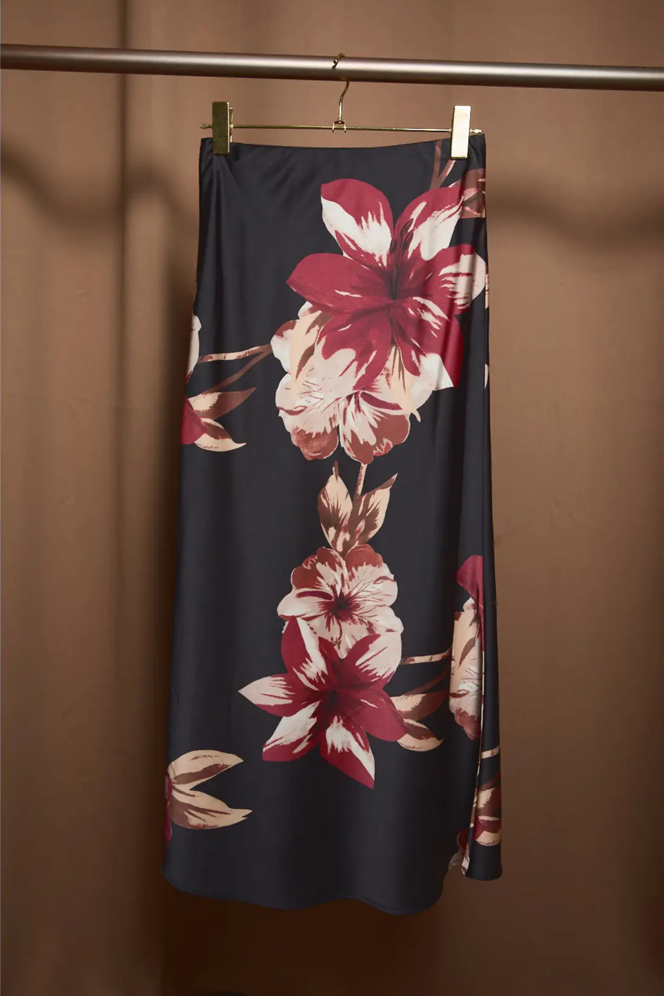 A dark navy midi skirt with a large red and cream floral print, hanging from a gold hanger on a clothing rack against a warm brown background.
