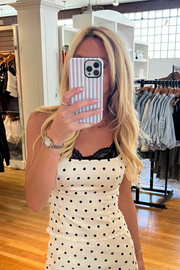 A woman takes a mirror selfie in a clothing boutique, wearing a white sleeveless dress with black polka dots and a black lace neckline. She is holding a striped phone case in a store with clothing racks and wooden floors visible in the background.