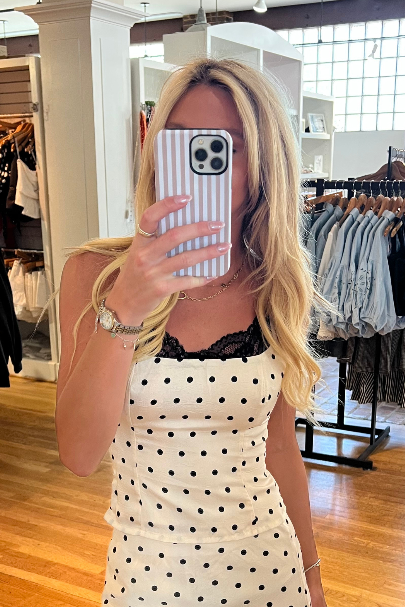 A woman takes a mirror selfie in a clothing boutique, wearing a white sleeveless dress with black polka dots and a black lace neckline. She is holding a striped phone case in a store with clothing racks and wooden floors visible in the background.