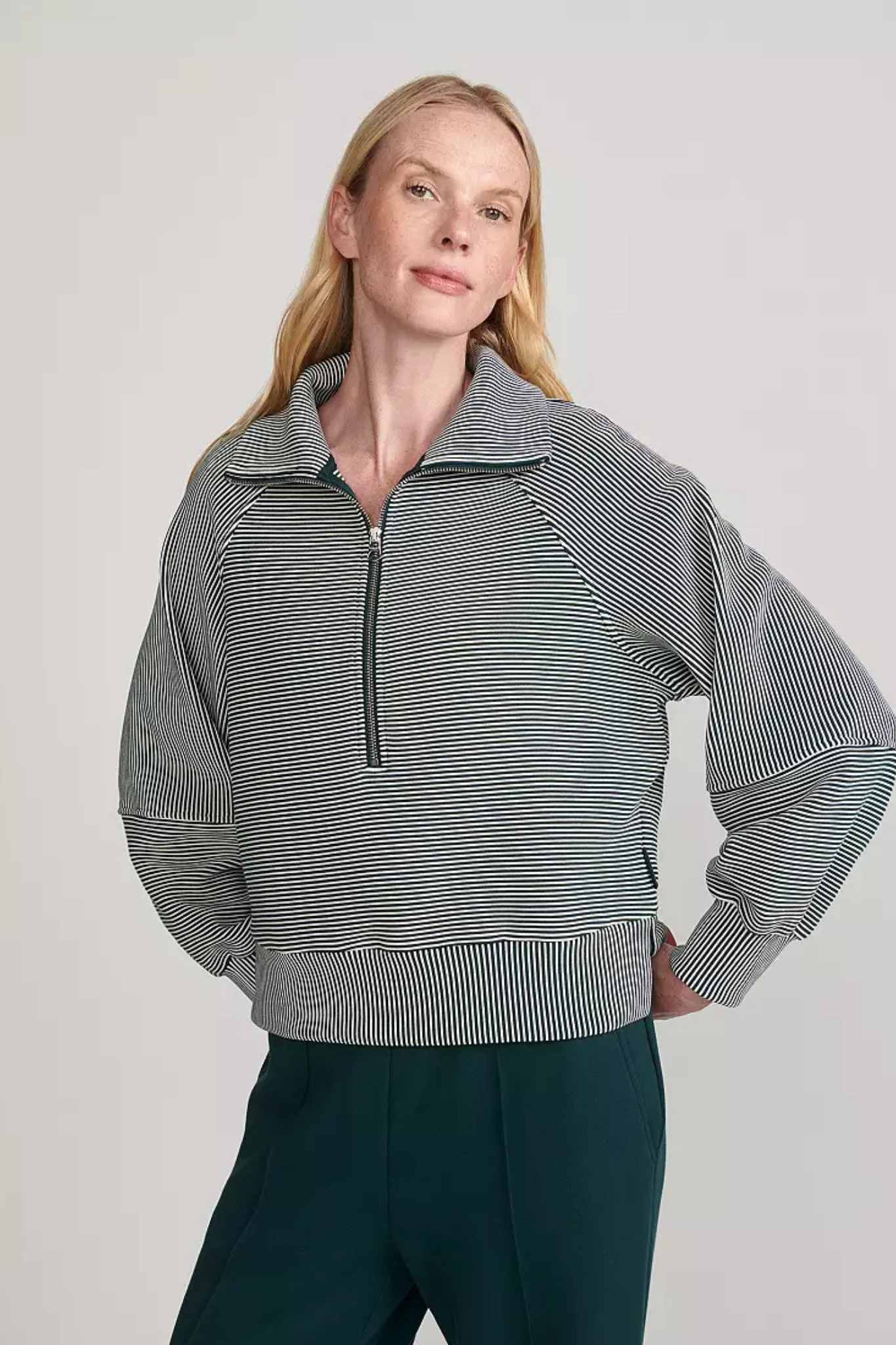 A woman modeling a gray and white vertically striped quarter-zip pullover top with a mock neck, paired with dark teal pants, against a light gray studio background.