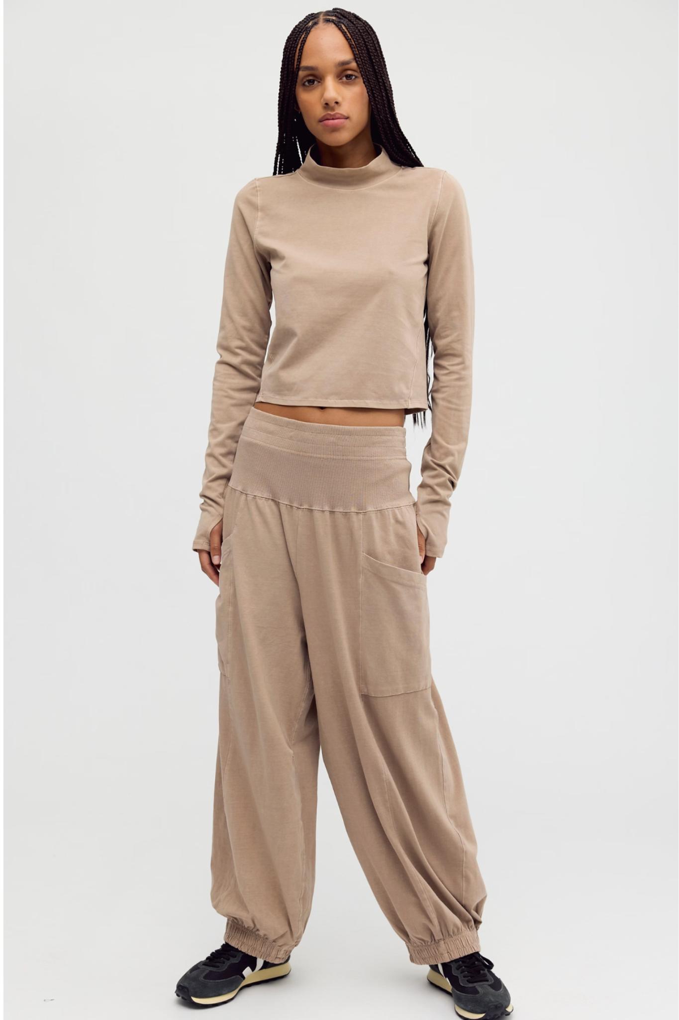 A model wearing a matching beige two-piece loungewear set, consisting of a long-sleeve cropped mock neck top and wide-leg jogger pants, paired with black sneakers, against a plain light gray background.