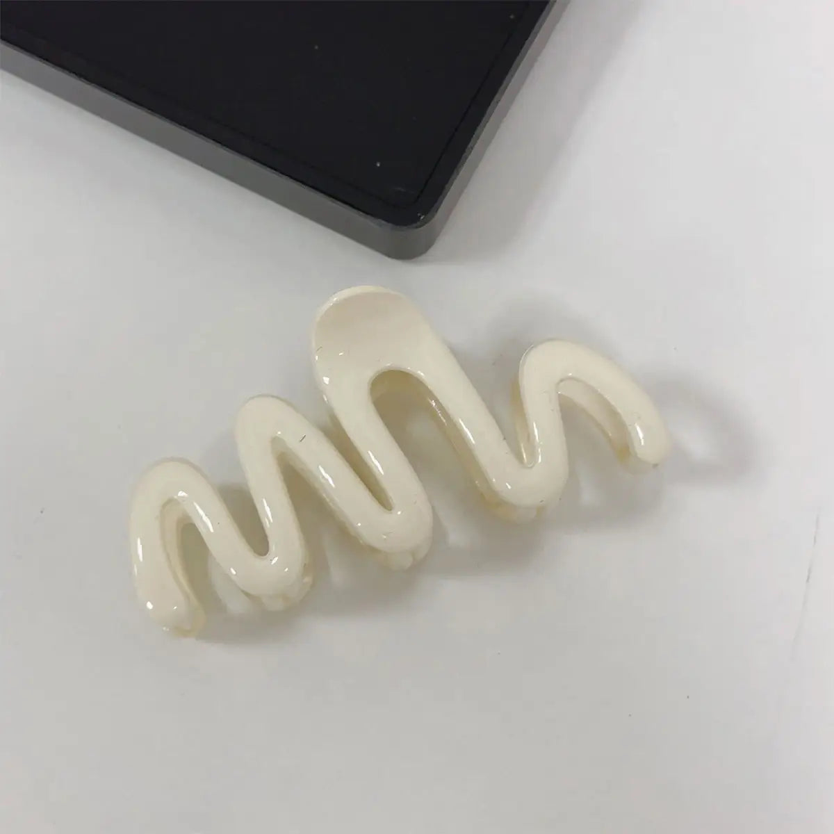 An off-white, glossy wavy hair claw clip lying on a light gray surface next to a black rectangular object.