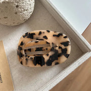 A tortoiseshell hair claw clip with a beige and black pattern, displayed on a textured beige fabric surface next to a decorative stone and a small card.
