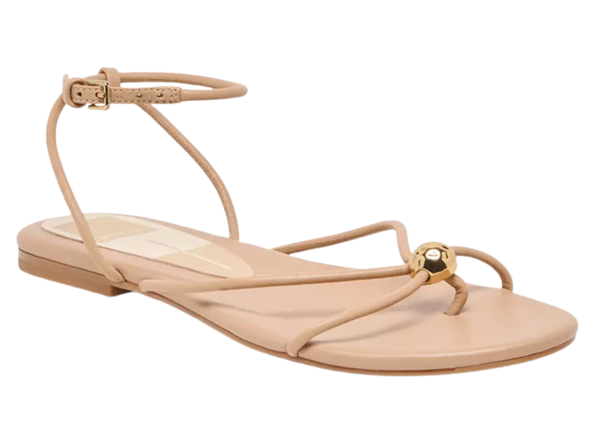 A single flat thong sandal in a light beige/nude leather with gold-tone hardware, featuring an ankle strap with a buckle and a decorative gold ball detail on the toe post, shown against a plain white background.