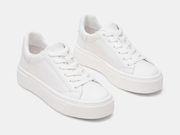 A pair of low-top platform sneakers in solid white, featuring white leather uppers, white laces, and thick white platform soles, shown on a light gray studio background.