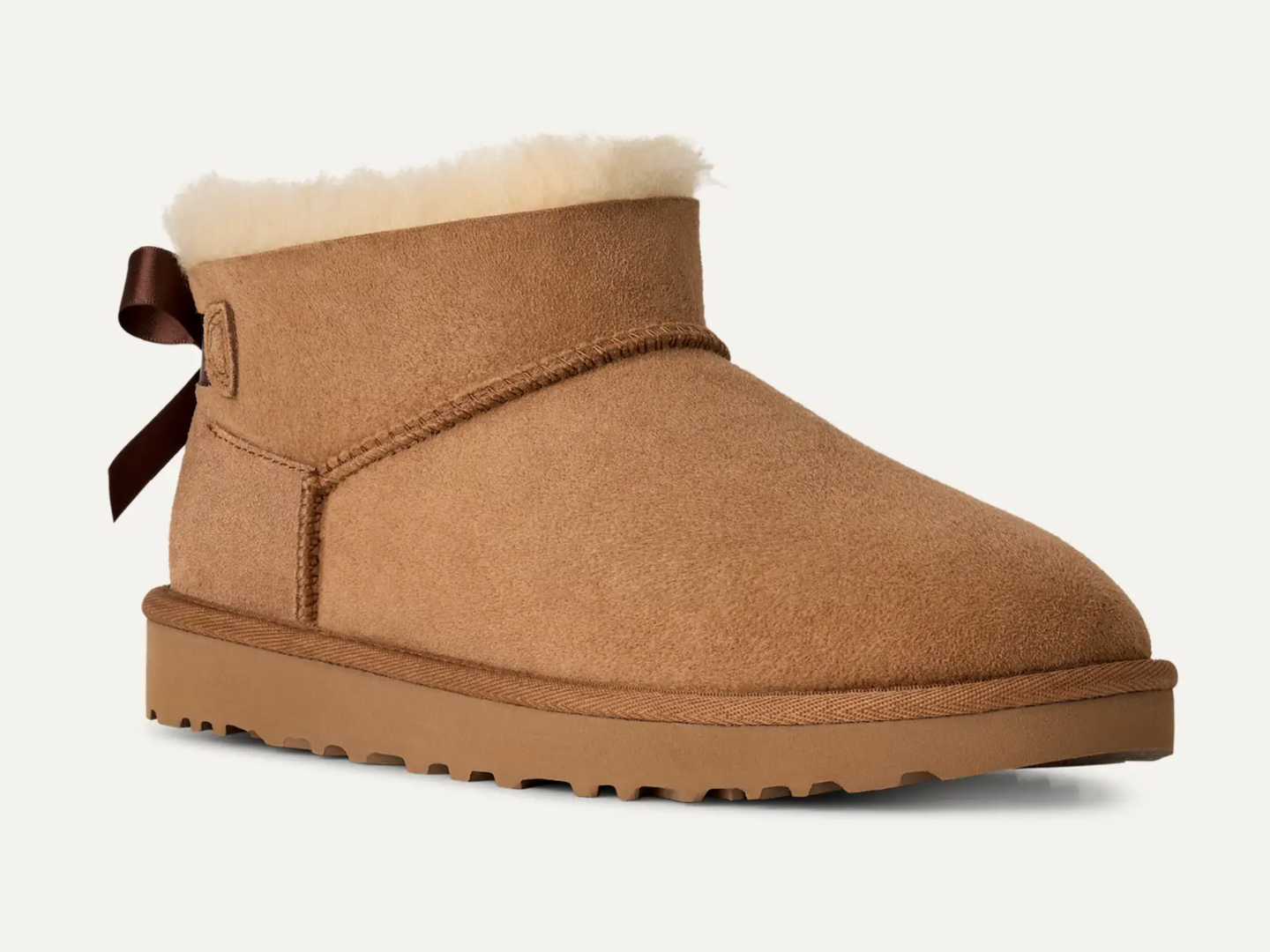 A classic low-top ankle boot in tan suede with a white shearling lining, a dark brown bow on the side, and a thick lug sole, shown against a plain off-white background.