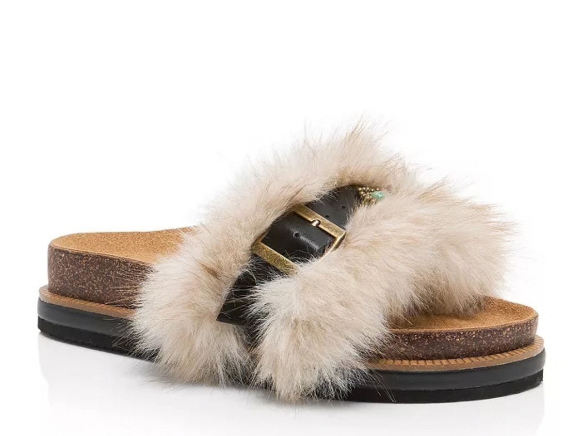 A single slide sandal featuring a thick, fluffy cream-colored fur strap with a black leather buckle and gold-tone metal hardware, set on a brown cork-look midsole with a black outsole, shown against a plain white background.