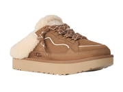 A UGG slip-on mule sneaker in tan suede with a thick cream shearling collar, brown and white patterned laces, and a beige platform sole.