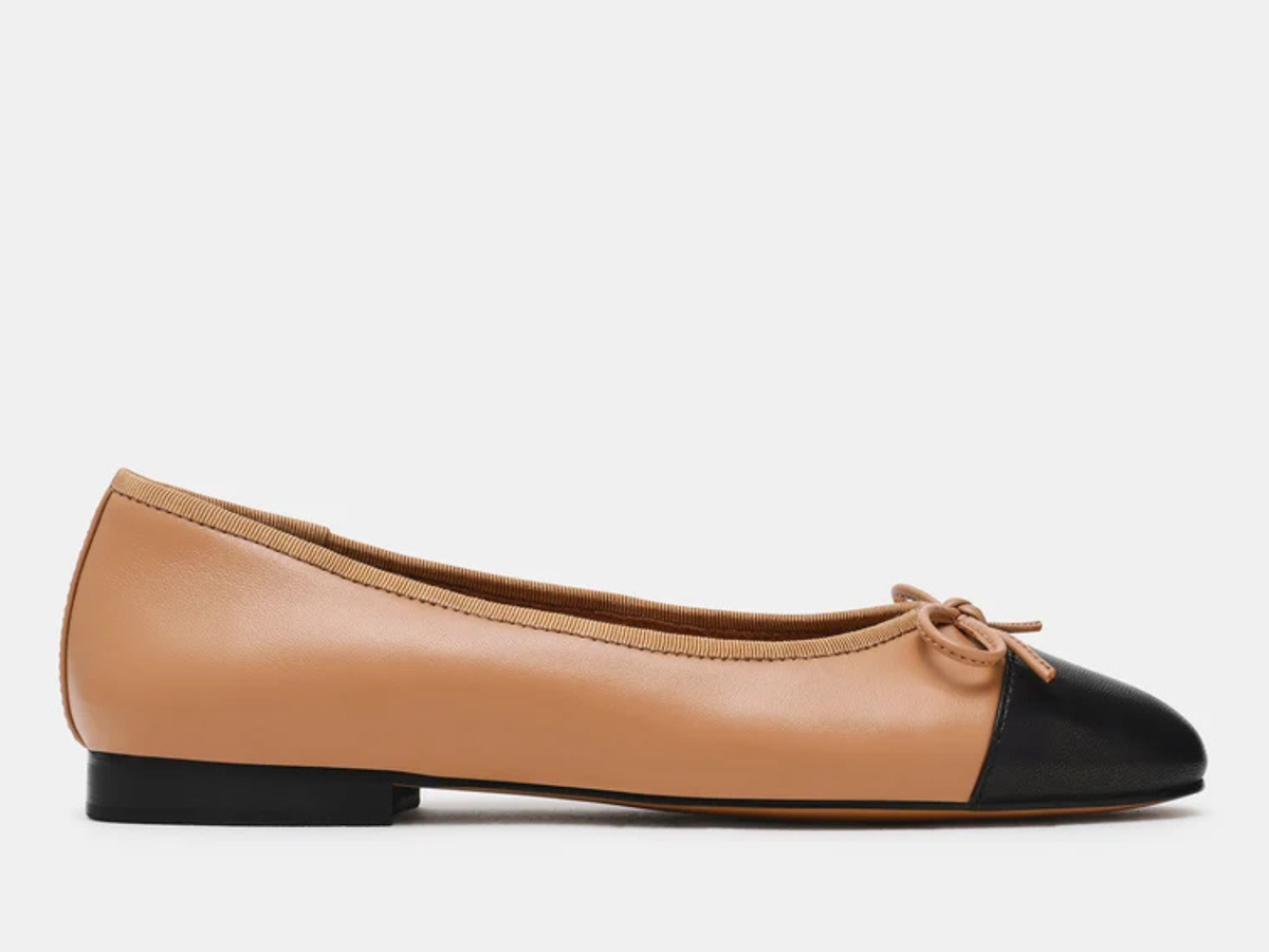A single ballet flat shoe in tan leather with a black pointed toe and black heel, featuring a matching tan bow on the front, shown against a plain light gray background.