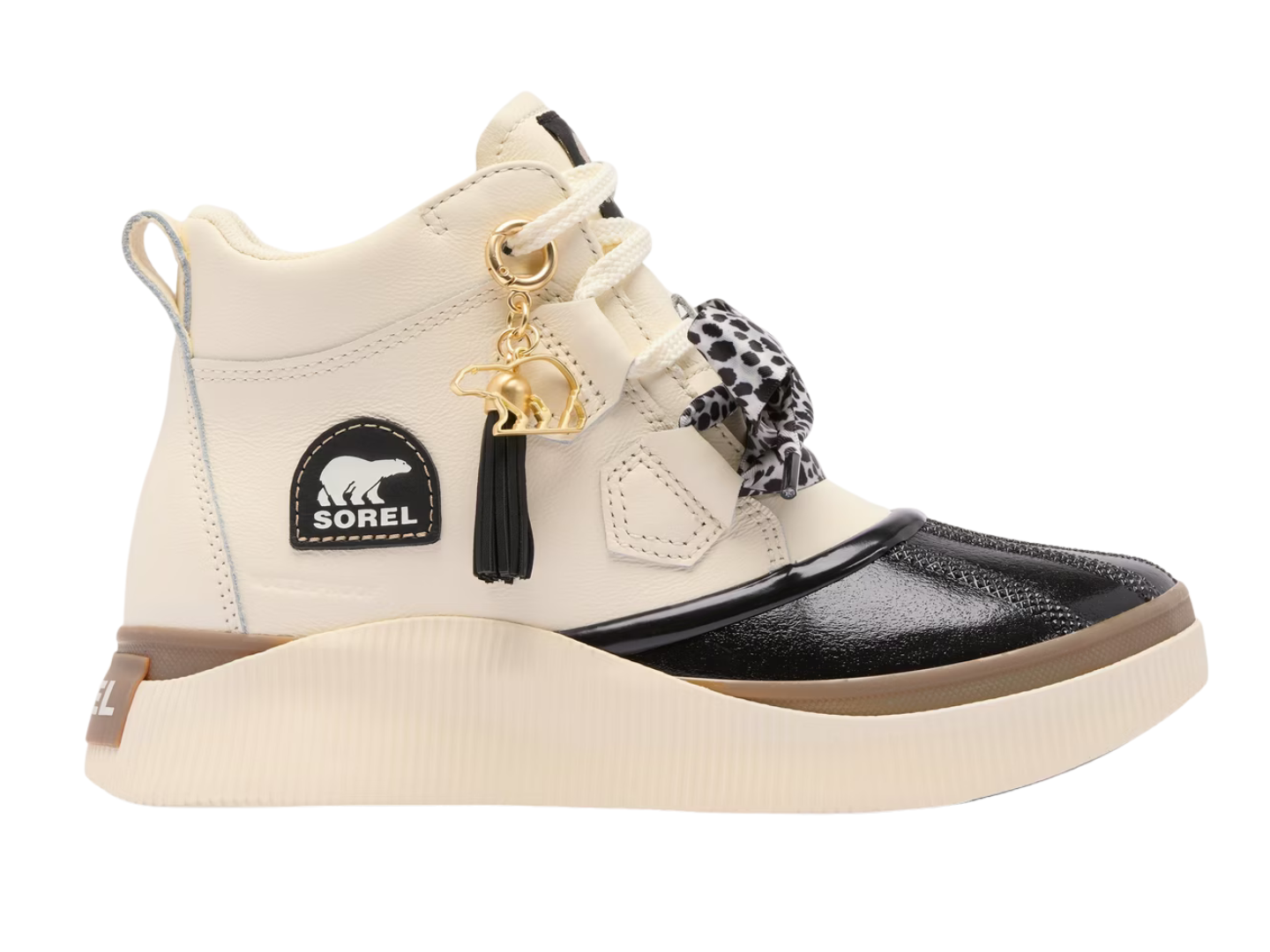 A Sorel mid-top sneaker in off-white and black, featuring a black patent leather toe cap, a cream rubber sole, a black Sorel logo patch on the side, and decorative gold-tone charms including a tassel and animal silhouette.