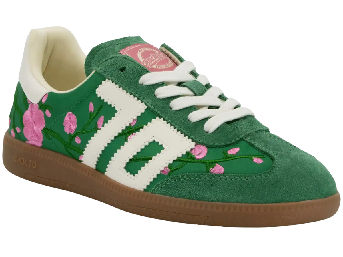 A green suede sneaker featuring a white serrated-style side stripe, white laces, a pink heart-shaped tongue label, and pink floral embroidery with green stems along the side. It has a brown gum sole.